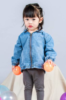 Stüdyoda, anaokulundaki küçük tatlı kız anaokulu çocuğunun tüm vücut fotoğrafları günlük sokakta kot ceket ve spor ayakkabıları giyer gri arka planda renkli plastik toplarla poz verir..