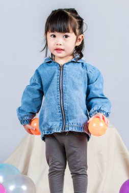 Stüdyoda, anaokulundaki küçük tatlı kız anaokulu çocuğunun tüm vücut fotoğrafları günlük sokakta kot ceket ve spor ayakkabıları giyer gri arka planda renkli plastik toplarla poz verir..