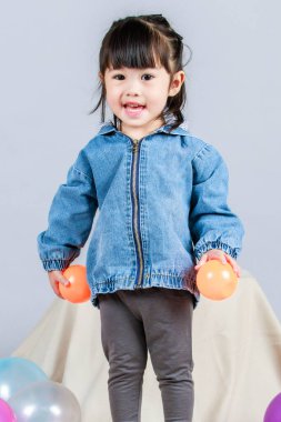 Stüdyoda, anaokulundaki küçük tatlı kız anaokulu çocuğunun tüm vücut fotoğrafları günlük sokakta kot ceket ve spor ayakkabıları giyer gri arka planda renkli plastik toplarla poz verir..