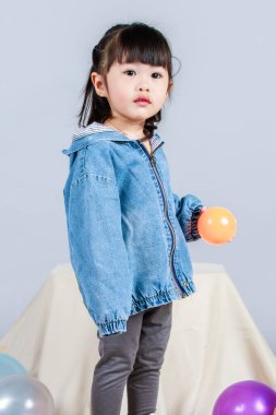 Stüdyoda, anaokulundaki küçük tatlı kız anaokulu çocuğunun tüm vücut fotoğrafları günlük sokakta kot ceket ve spor ayakkabıları giyer gri arka planda renkli plastik toplarla poz verir..