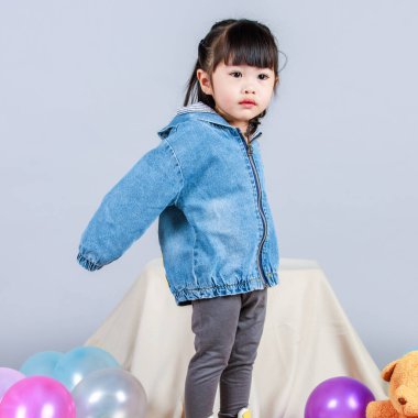 Stüdyoda, anaokulundaki küçük tatlı kız anaokulu çocuğunun tüm vücut fotoğrafları günlük sokakta kot ceket ve spor ayakkabıları giyer gri arka planda renkli plastik toplarla poz verir..