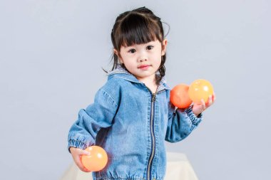 Stüdyoda, anaokulundaki küçük tatlı kız anaokulu çocuğunun tüm vücut fotoğrafları günlük sokakta kot ceket ve spor ayakkabıları giyer gri arka planda renkli plastik toplarla poz verir..