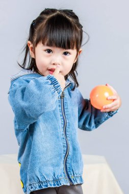 Stüdyoda, anaokulundaki küçük tatlı kız anaokulu çocuğunun tüm vücut fotoğrafları günlük sokakta kot ceket ve spor ayakkabıları giyer gri arka planda renkli plastik toplarla poz verir..