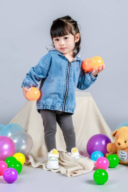 Stüdyoda, anaokulundaki küçük tatlı kız anaokulu çocuğunun tüm vücut fotoğrafları günlük sokakta kot ceket ve spor ayakkabıları giyer gri arka planda renkli plastik toplarla poz verir..