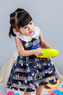 Küçük, masum anaokulu anaokulu kızı, mavi bir elbise giymiş, gri arka planda renkli balonlar tutan kameraya gülümseyen bir kızın portresi..