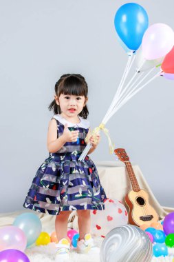 Küçük, masum anaokulu anaokulu kızı, mavi bir elbise giymiş, gri arka planda renkli balonlar tutan kameraya gülümseyen bir kızın portresi..