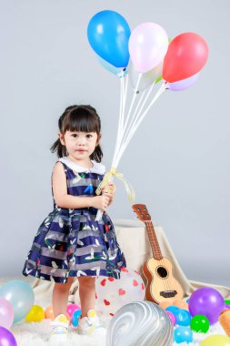 Küçük, masum anaokulu anaokulu kızı, mavi bir elbise giymiş, gri arka planda renkli balonlar tutan kameraya gülümseyen bir kızın portresi..