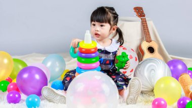 Renkli helyum balonları arasında oturmuş gri arka planda plastik halkalar ve dinozor oynayan küçük, masum anaokulu anaokulu kızı kızı fotoğrafı..