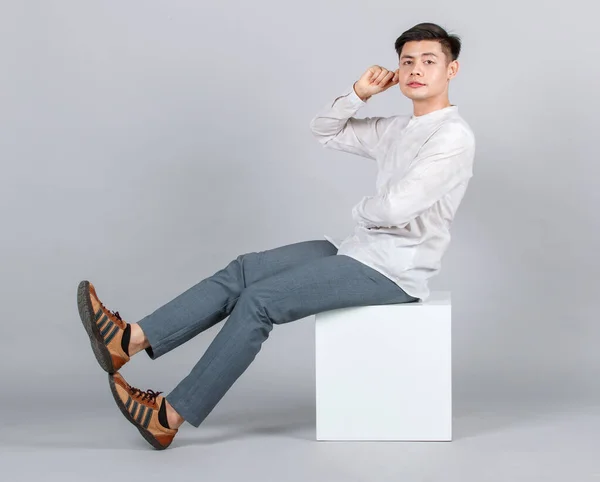 スタジオショットの千年のアジアのハンサムな若い男性モデルでカジュアル服に座って上の白い正方形のスツール見てカメラで灰色の背景 — ストック写真