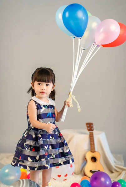小可爱的幼稚园小女孩穿着休闲长衫站在那里笑着看着镜头拿着色彩斑斓的气球灰色背景的画像 — 图库照片