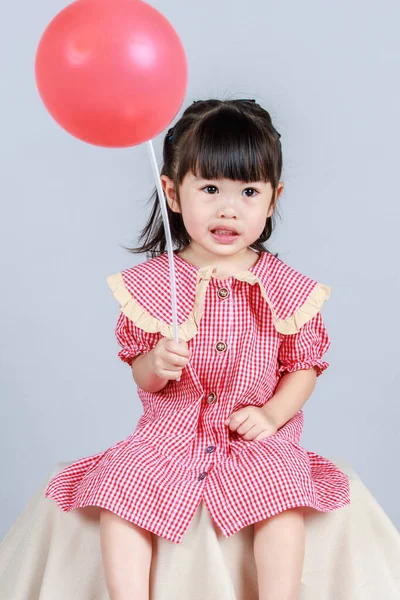 演播室特写小可爱的小女孩 身穿红色长裙 独自一人坐在台布上 手持氦气气球棒 在灰色的背景下微笑 — 图库照片