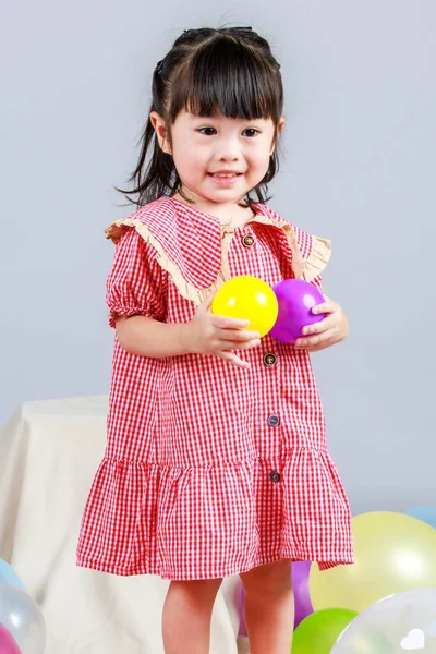 Küçük şirin anaokulu çocuğu, kırmızı uzun elbiseli, tek başına gülümseyen, gri arka planda renkli helyum hava partisi balonlarıyla oynayan kız çocuğunun portresi.