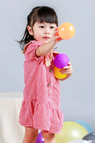 Küçük şirin anaokulu çocuğu, kırmızı uzun elbiseli, tek başına gülümseyen, gri arka planda renkli helyum hava partisi balonlarıyla oynayan kız çocuğunun portresi.