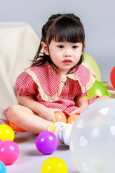 Küçük şirin anaokulu çocuğu, kırmızı uzun elbiseli, gülümseyen, gri arka planda renkli helyum hava balonlarıyla oynayan kız çocuğunun portresi..