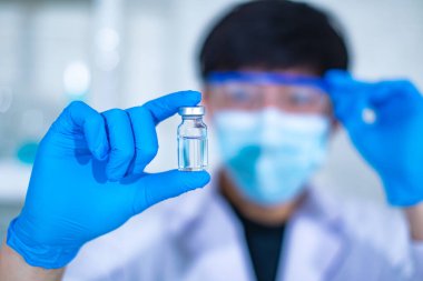 Asyalı, profesyonel, beyaz laboratuvar önlüklü, yüz maskeli ve güvenlik gözlüklü bir bilim adamının aşı cam şişesini bulanık ön planda tutarken çekilmiş portresi..