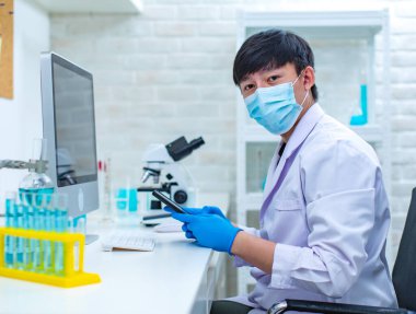 Beyaz laboratuvar önlüğü ve güvenlik maskesi takmış Asyalı profesyonel erkek bilim adamı klavye bilgisayarında oturmuş deney bilgilerini analiz ediyor ve hastane laboratuarındaki çalışma masasında çalışıyor..