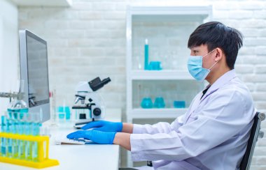 Beyaz laboratuvar önlüğü ve güvenlik maskesi takmış Asyalı profesyonel erkek bilim adamı klavye bilgisayarında oturmuş deney bilgilerini analiz ediyor ve hastane laboratuarındaki çalışma masasında çalışıyor..