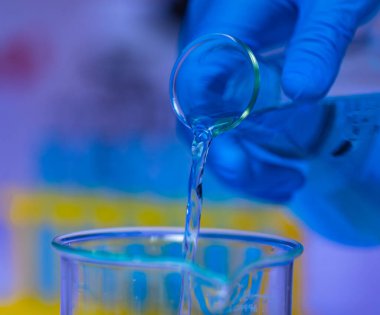 Yakın çekim stüdyo çekiminde Erlenmeyer şişesinden cam şişeye dökülen mavi solüsyon reaksiyonlu sıvı numunesi bilim adamı eliyle test tüpünün arka planında bulunan plastik eldiven takıyor..