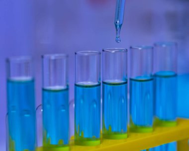 Laboratuvar cam damlatıcısının, laboratuvar laboratuarındaki bilimsel deneyleri analiz etmek için raftaki cam test tüpüne mavi reaksiyonlu çözücü sıvı numunesi attığı yakın çekim..