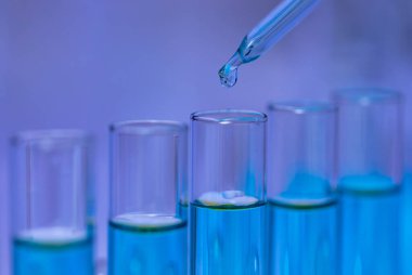 Laboratuvar cam damlatıcısının, laboratuvar laboratuarındaki bilimsel deneyleri analiz etmek için raftaki cam test tüpüne mavi reaksiyonlu çözücü sıvı numunesi attığı yakın çekim..