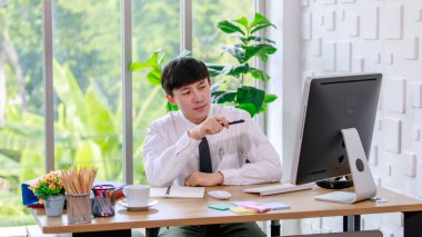 Asyalı profesyonel iş adamının resmi gömlekli ve kravatlı portre çekimi. Çalışma masasında bilgisayar monitörü fare ve kırtasiye malzemesi ile kameraya bakıyor..