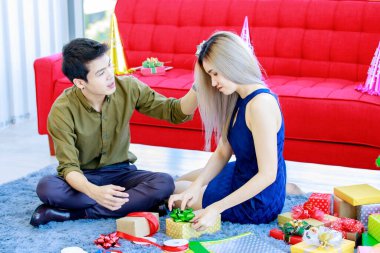 Milenyum genç Asyalı erkek arkadaş oturma odasında halı döşemesinde oturmuş yeni yılı paketleyen ve hediye kutularını kağıt ve kurdeleyle dolduran kız arkadaşını okşuyor..