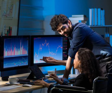 Beyaz erkek hisse senedi yatırımcısı Afrika kökenli Amerikalı kadın iş kadınıyla ticaret odasında oturmuş grafik analiz ekranları ve monitörlerle konuşuyor..