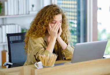 Kıvırcık saç stili mutsuz genç bunalımda yorgun, uykusuz, Kafkasyalı hamile, rahat giyinmiş, iş yerinde dizüstü bilgisayarla çalışan, baş ağrısı çeken bir anne..