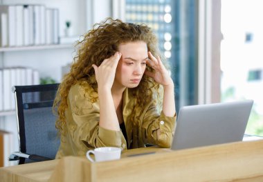 Kıvırcık saç stili mutsuz genç bunalımda yorgun, uykusuz, Kafkasyalı hamile, rahat giyinmiş, iş yerinde dizüstü bilgisayarla çalışan, baş ağrısı çeken bir anne..