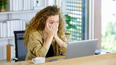 Kıvırcık saç stili mutsuz genç bunalımda yorgun, uykusuz, Kafkasyalı hamile, rahat giyinmiş, iş yerinde dizüstü bilgisayarla çalışan, baş ağrısı çeken bir anne..