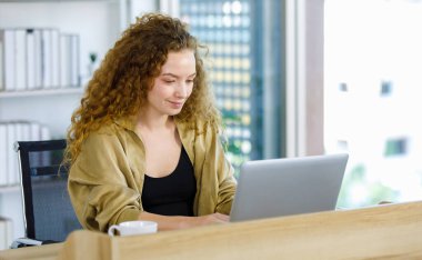 Kıvırcık saç stili, mutlu, sağlıklı, beyaz, hamile, iş kadını, günlük kıyafetli, iş masasında dizüstü bilgisayarla oturuyor.