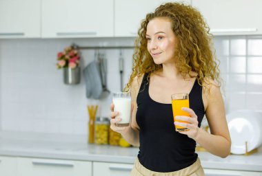 Mutlu kıvırcık saç stili Kafkas hamile anne gündelik kıyafetleri içinde elinde bir bardak portakal suyu ve sütü evdeki mobilyalı mutfakta kahvaltıda seçiyor..