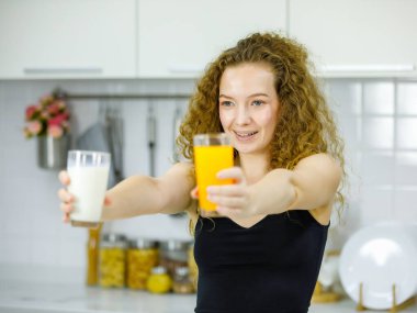 Mutlu kıvırcık saç stili Kafkas hamile anne gündelik kıyafetleri içinde elinde bir bardak portakal suyu ve sütü evdeki mobilyalı mutfakta kahvaltıda seçiyor..