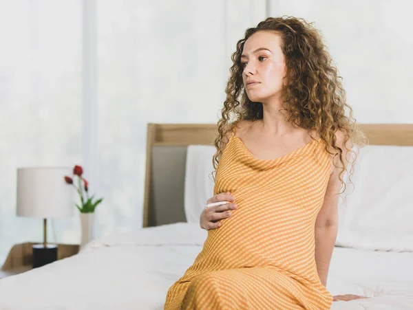 Curly hairstyle young unhappy unhealthy bad behavior Caucasian pregnancy mother in maternity long dress cloth sit on bed in bedroom holding smoking cigarette taking risk and danger to unborn child.