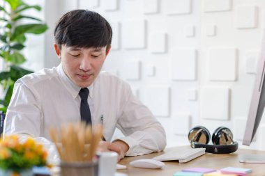 Asyalı profesyonel iş adamının resmi gömlekli ve kravatlı portre çekimi. Çalışma masasında bilgisayar monitörü fare ve kırtasiye malzemesi ile kameraya bakıyor..