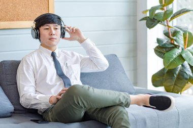 Resmi iş gömleği ve kravatlı Asyalı genç, yakışıklı, profesyonel iş adamı rahat koltukta oturmuş akıllı telefonlu internet üzerinden müzik dinliyor..