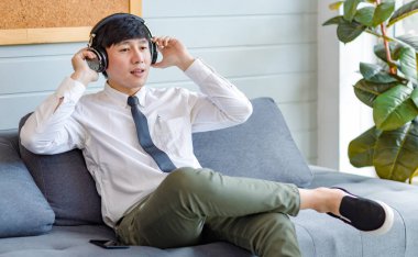 Resmi iş gömleği ve kravatlı Asyalı genç, yakışıklı, profesyonel iş adamı rahat koltukta oturmuş akıllı telefonlu internet üzerinden müzik dinliyor..