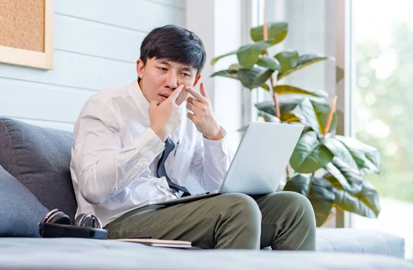 Asian young male businessman employee in formal business outfit sitting on cozy sofa couch with laptop holding hands and fingers squeezing inflammatory acne pimple on cheekbone in living room at home.