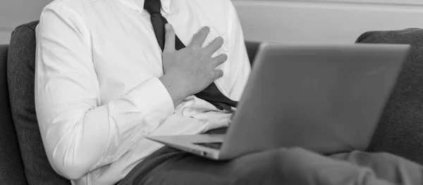 身着正式商务服装的不明身份男性生意人坐在舒适的沙发上 手牵着手提电脑 左胸紧握着患有心脏病的单色黑白照片 — 图库照片