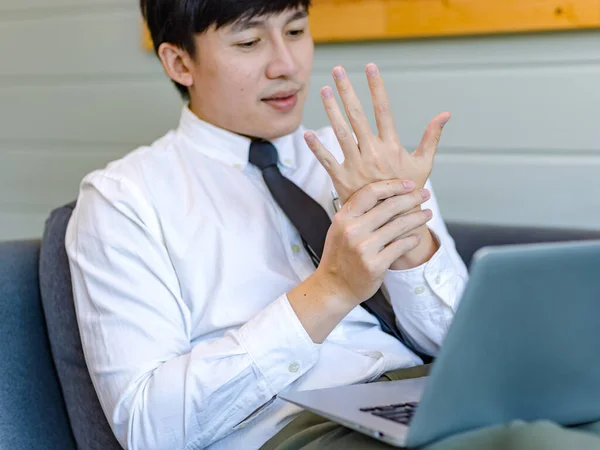 Asian tired overwork male businessman employee in business outfit sitting on sofa working with laptop computer holding hand rubbing massage on stiff injury neck and shoulder from office syndrome.