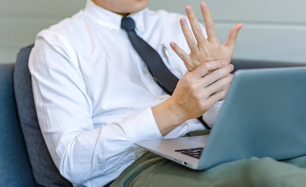 Asian tired overwork male businessman employee in business outfit sitting on sofa working with laptop computer holding hand rubbing massage on stiff injury neck and shoulder from office syndrome.