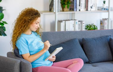 Beyaz, mutlu, uzun kıvırcık saçlı, sağlıklı hamile, günlük kıyafetli, gülümseyen, elinde kalemle oturan anne oturma odasında yeni doğan çocuk için not defteri hazırlıyor..