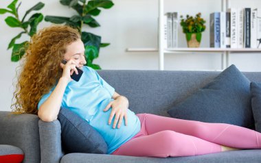 Beyaz, güzel, uzun kıvırcık saçlı sağlıklı hamile anne rahat bir şekilde koltuğa oturup karnına dokunuyor oturma odasında akıllı telefonla konuşuyor..