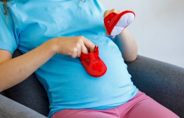 Beyaz, mutlu, uzun kıvırcık saçlı, sağlıklı hamilelik annesi rahat elbiseler içinde, rahat koltukta oturmuş, kucağında bebek kırmızı ayakkabılarına bakıyor ve yeni doğmuş bir çocuk bekliyor..