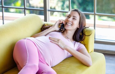 Milenyum Kafkasyalı genç, mutlu, sağlıklı, koca göbekli hamile anne rahat bir kanepede uzanmış gülümsüyor akıllı telefonla konuşurken karnına dokunuyor..