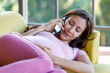 Milenyum Kafkasyalı genç, mutlu, sağlıklı, koca göbekli hamile anne rahat bir kanepede uzanmış gülümsüyor akıllı telefonla konuşurken karnına dokunuyor..