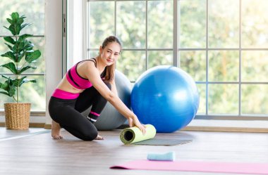 Asian young healthy slim beautiful female sporty model in crop top sportswear sitting smiling rolling yoga mat on floor in glass windows fitness gym aerobic classroom with pilates exercise balls.