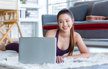 Millennial Asian young healthy slim strong sexy female sporty model in casual crop top sportswear laying down smiling on carpet floor in living room learning yoga workout class via laptop computer.
