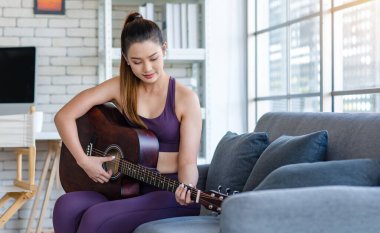Milenyum Asyalı genç, sağlıklı, güzel, sağlıklı, güçlü, seksi, spor model, rahat giyinmiş, güler yüzlü, evdeki oturma odasındaki rahat koltukta akustik gitar çalan kadın..