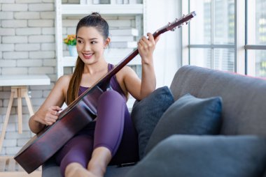 Milenyum Asyalı genç, sağlıklı, güzel, sağlıklı, güçlü, seksi, spor model, rahat giyinmiş, güler yüzlü, evdeki oturma odasındaki rahat koltukta akustik gitar çalan kadın..